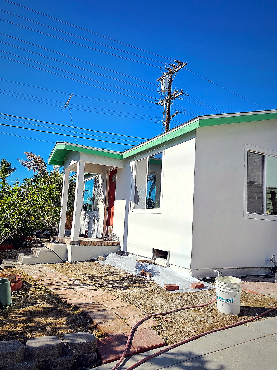 Front of the re-stuccoed home — fresh smooth white walls, original green eaves and red entry door preserved, paver walkway leading to the front porch.